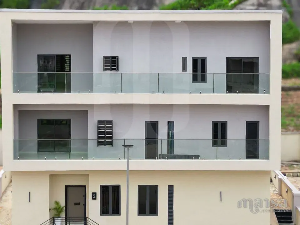 The completed 5-bedroom standalone house in Katampe Extension, Abuja, seen from the front, with glass-balustraded balconies on two floors and the Katampe rock outcrop and green hillside behind it